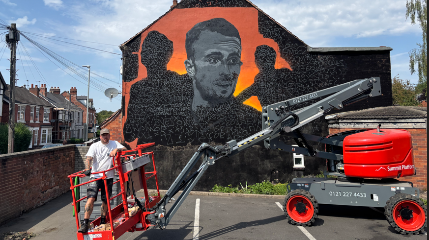 A man painting a black and orange mural. The mural shows a painting of a man looking to the right in black and white on an orange background. The artist, a man, wears a baseball cap and stands in the red canopy of a machine to raise him up in the air.