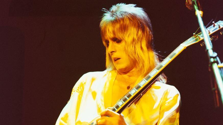 Mick Ronson has a blonde shoulder length mullet-style volumed hair and is holding a guitar whilst performing on stage. There is a yellow light over him and he has his eyes closed and mouth slightly open. He appears to be wearing black eyeshadow.