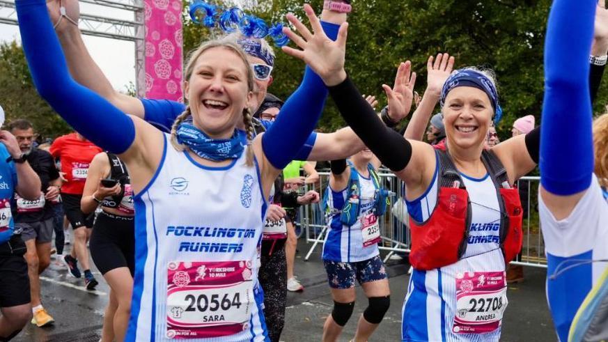 Two smiling women wearing white and blue Pocklington Runners tops raise their hands in the air as they start the Yorkshire Marathon among a crowd of other runners. 