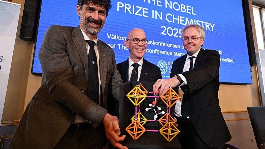 Three members of the Nobel committee hold a model of metal-organic frameworks, which is an orange, pink and yellow structure.
