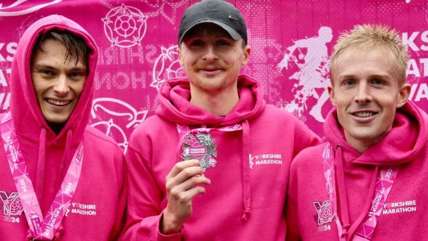 The winner of the men's race shows off his medal while flanked by the runners who finished second and third. All three are wearing pink hoodies and smiling. They are standing in front of a pink Yorkshire Marathon banner. 