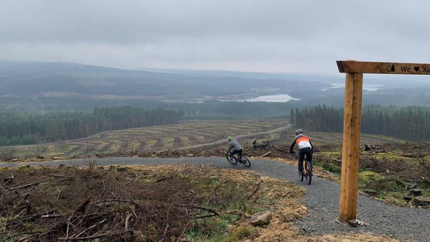 Two cyclists descend on mountain bikes along a gravel path which winds into the valley. In the distance you can see Kielder Reservoir.