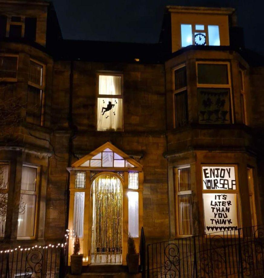 A house with gold fringe over the door, fairy lights over the gate and the lyrics "Enjoy yourself, it's later than you think" in the window