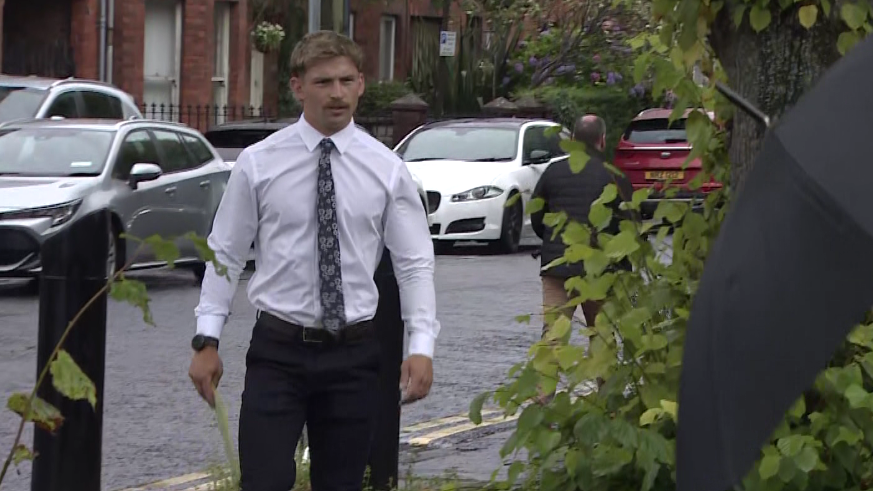 Kavan Keenan is a young man with short blonde/ brown hair and a moustache. He is walking towards the gates into the court as the picture is taken. He is wearing a white long sleeve shirt and a black patterned tie. Behind him is a silver car and two white cars parked along the street in front of red brick terrace houses.
