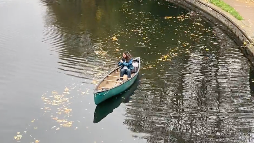 Chloe Edwards is sat paddling down Regents Canal in the Community Canoe. The Canoe is green and centre-frame in the shot.