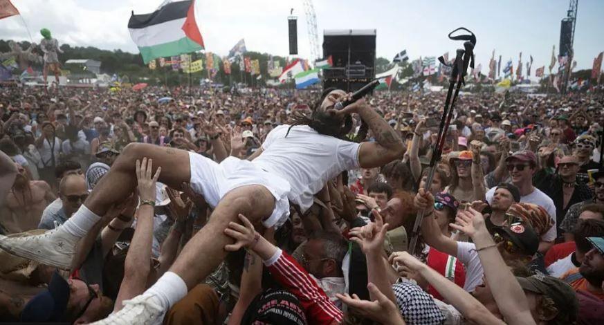 Bobby Vylan, lead singer of Bob Vylan, is held aloft by fans as he lies out. at the front of the band's show at Glastonbury Festival 2025, on Saturday. He has a microphone to his mouth and is wearing white T-shirt and shorts with white socks and hi-top sneakers.