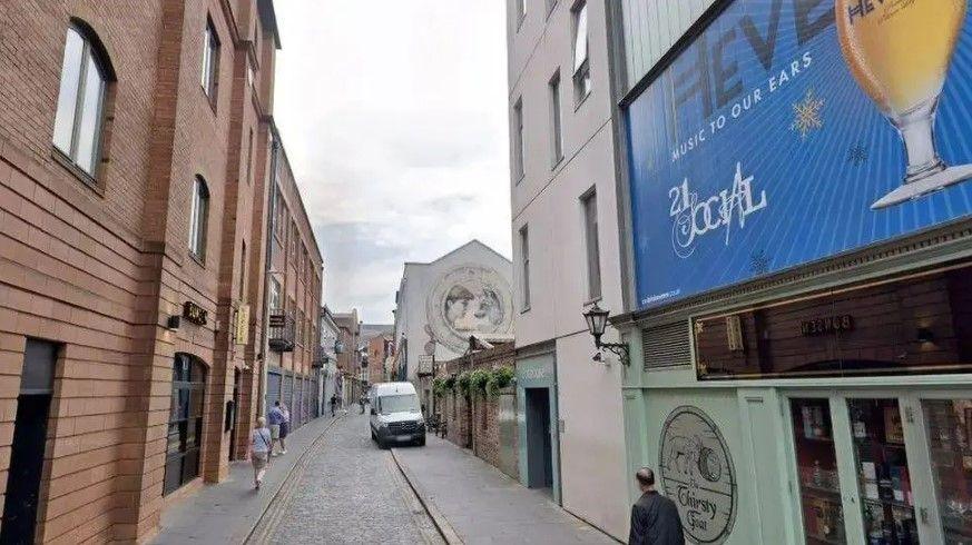 A small street with grey cobblestones and paving. On one side is a red brick building with black shutters. On the other is a number of businesses, the one closest to the camera has a light green front with a large blue billboard.