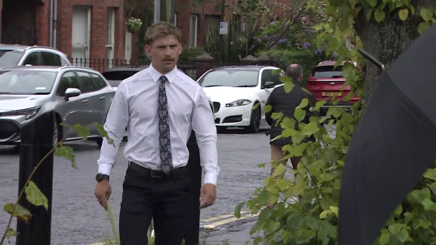 Kavan Keenan is a young man with short blonde/ brown hair and a moustache. He is walking towards the gates into the court as the picture is taken. He is wearing a white long sleeve shirt and a black patterned tie. Behind him is a silver car and two white cars parked along the street in front of red brick terrace houses.