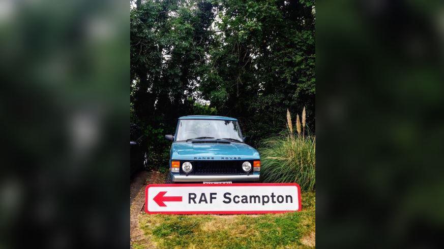 The RAF Scampton sign with a red arrow pointing left in front of a blue Range Rover to show how big it is