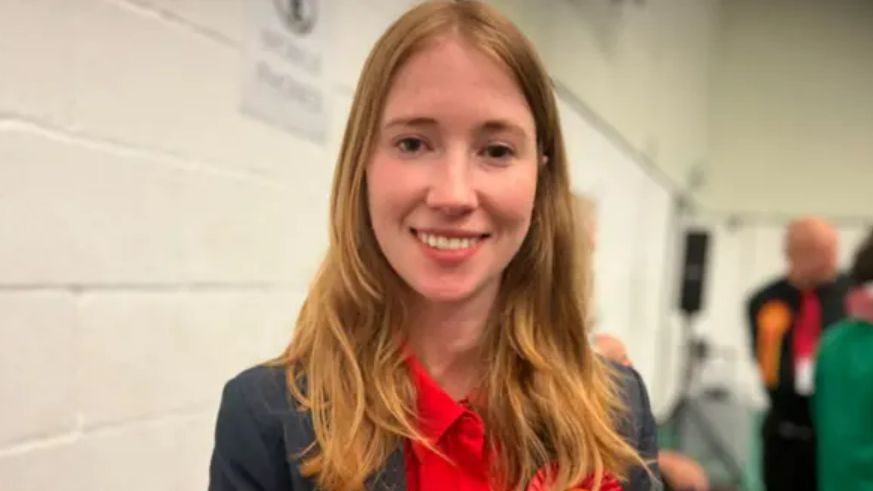 Helena Dollimore, the Labour MP for Hastings and Rye, wears a red shirt and red Labour rosette and smiles at the camera. She appears to be at an election count. 