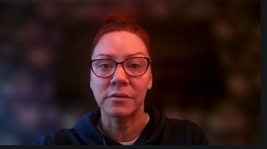 A woman stares at the camera with glasses on and her hair tied back and is wearing a dark-coloured top.