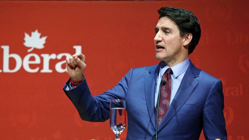 Justin Trudeau standing at a podium in front of a red back drop. He is wearing a blue suit and patterned tie and is pointing with one raised hand