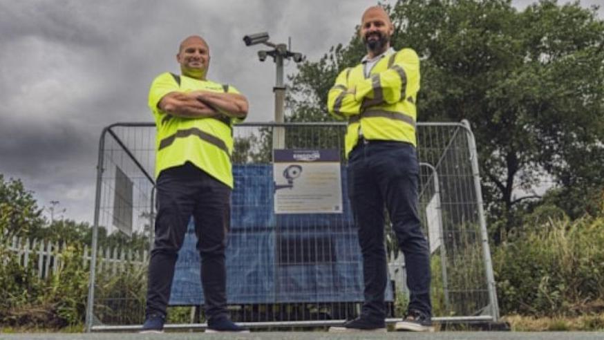 Two men stand posing for the camera with their arms folded, wearing hi-vis jackets, jeans and trainers. They are stood in front of the cameras, which have metal fences around them.