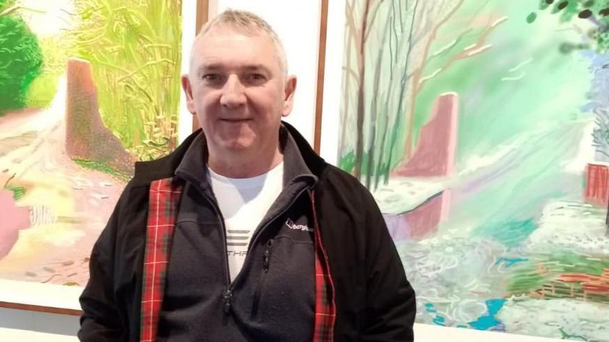 Andrew Lodge - a smiling man of around 60. He has short grey hair and is wearing a black coat, dark-coloured tracksuit top and a white shirt. He is standing between two large watercolour paintings