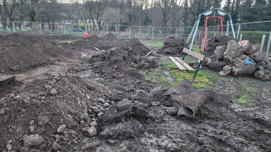 Lots of earth is piled up with big boulders next to a dirty blue and red swing. The playpark looks like a construction site and is surrounded by metal fencing