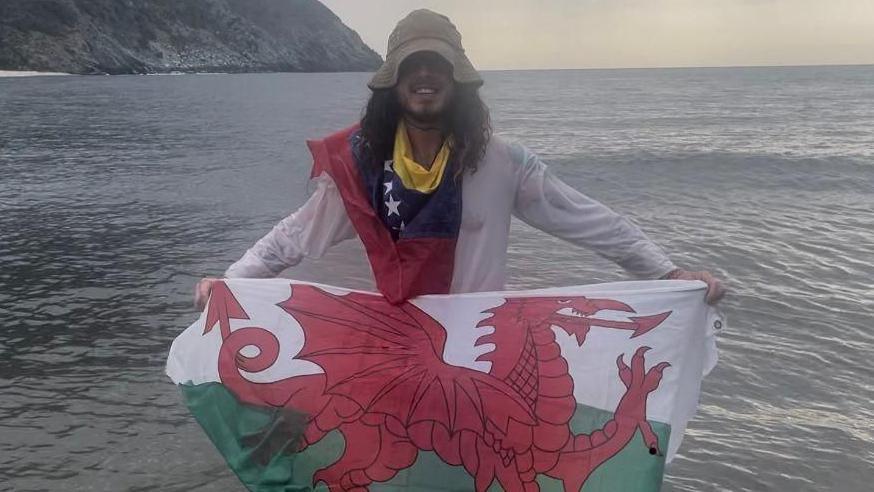Yn y llun yma mae Ollie yn cario baner Cymru ar lan y mor. Mae e'n gwisgo siwmper hufen a chap hufen hefyd. Mae ganddo wallt brown hir.