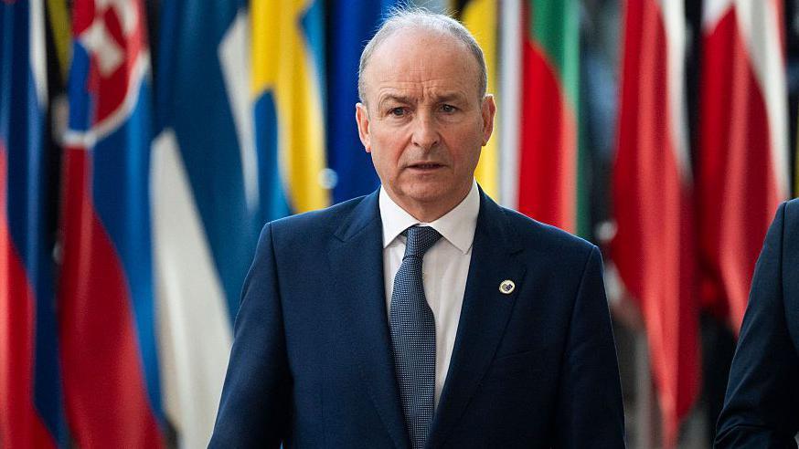 A man in a navy suit wants in front of a number of international flags. He is wearing a white shirt and navy tie.