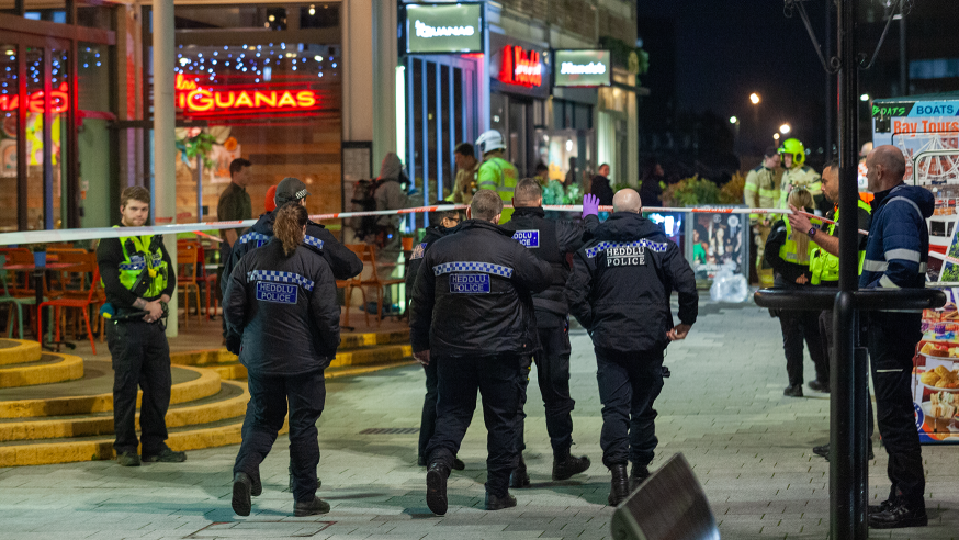 Police officers near Las Iguanas restaurant in Cardiff Bay after the incident, with police tape stretched across the pavement and firefighters in the background