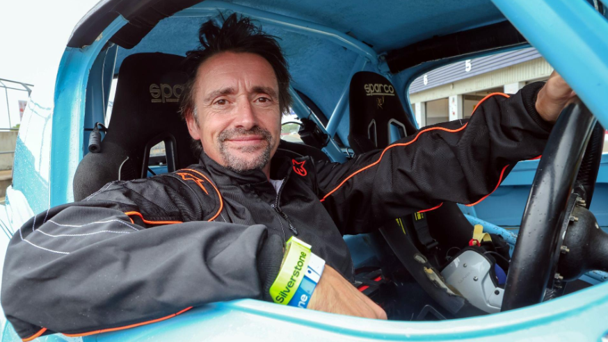 Richard Hammond sat in a light blue car holding the steering wheel. He is looking at the camera and smiling.