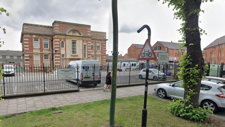 Google view of the red-bricked building with mullioned windows. There is a car park in the foreground and a sign at the entrance.