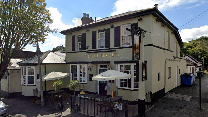 The exterior of The Three Colts pub, which is a two-storey, cream coloured building. It has two benches outside, both with umbrellas and enclosed within a black, metal perimeter fence. The building has a door and two windows on the bottom floor, with three windows on the first floor. It is situated on a residential street.