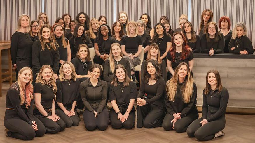 A large group of women in a room smiling at the camera. They are all wearing black and some are kneeling down, while others are sitting or standing.