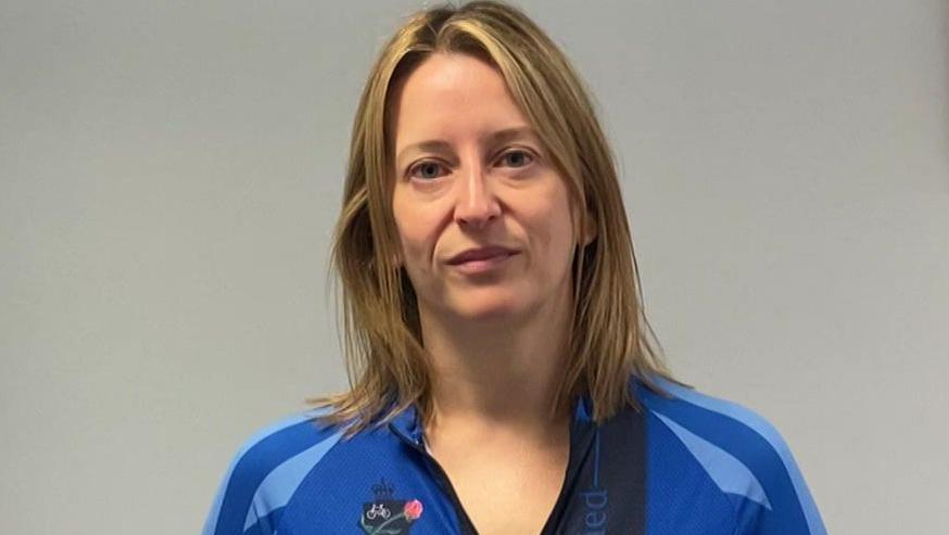 A blonde woman with shoulder length hair stands in front of a grey wall. She looks serious, and is wearing a blue long sleeved cycling top with UK Police Unity Tour written on it.