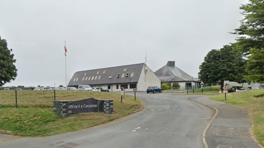 A screenshot from Google Maps showing the primary school, Ysgol y Creuddyn. Looking at the white building from the point of view of the main road. A Welsh flag hangs on a pole in the distance. Trees either side of the school.