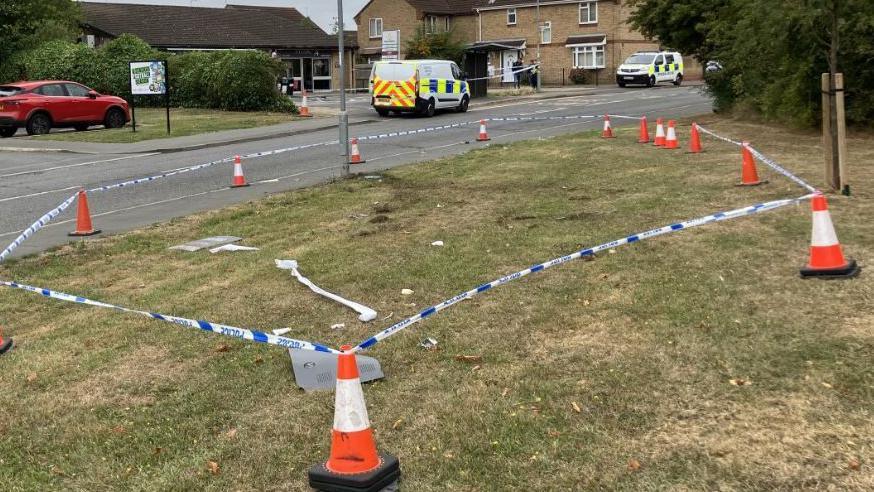 Police tape and orange and white cones around a section of grass verge with debris inside it - with the damaged Morrisons in the background along with two police vans