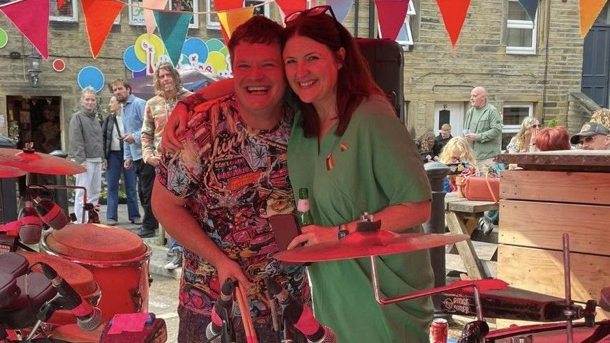 Hilary Bowyer (R) wearing a green dress and with her arm around Bongo Dave (L), who is wearing a patterned t-shirt at the event. They are stood next to musical instruments and people are stood looking at them behind them.