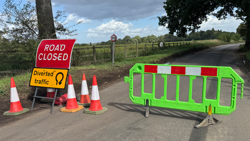 Playford Road in Suffolk blocked off by a bright green barrier, traffic cones, and a Road Closed sign. 