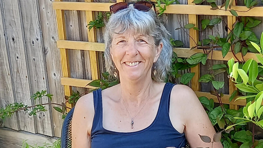 Jan sat in front of a garden fence. She has short grey hair and wears a navy vest, a necklace and brown sunglasses on her head. She is smiling.
