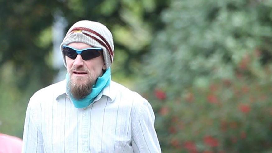 A man wearing a striped shirt and and hat while carrying sunglasses.