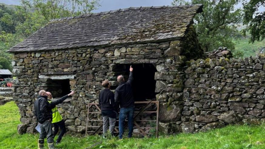 A stone barn in a field. Four men are looking at the building and pointing at it.