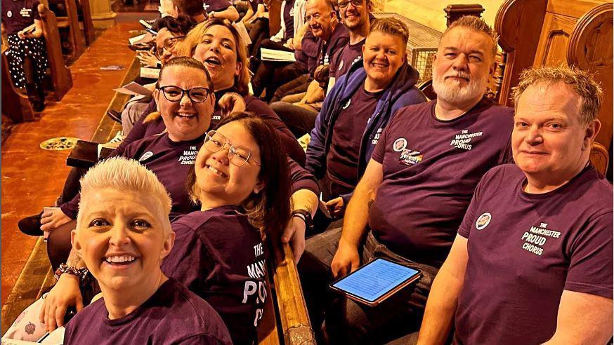 Members of the Manchester Proud Chorus choir pose for a photo wearing pink polo shirts emblazoned with the choir's name while sat on pews at a church.