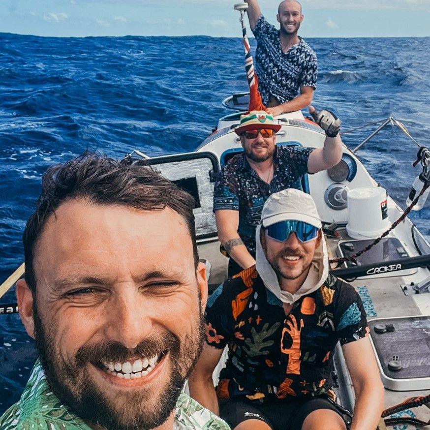 The man with a beard in the foreground is smiling and wearing a green top. The next two men have sunglasses on and the man at the back has his right arm up near a pole that has a flag on it. The sea is behind the boat they are in.
