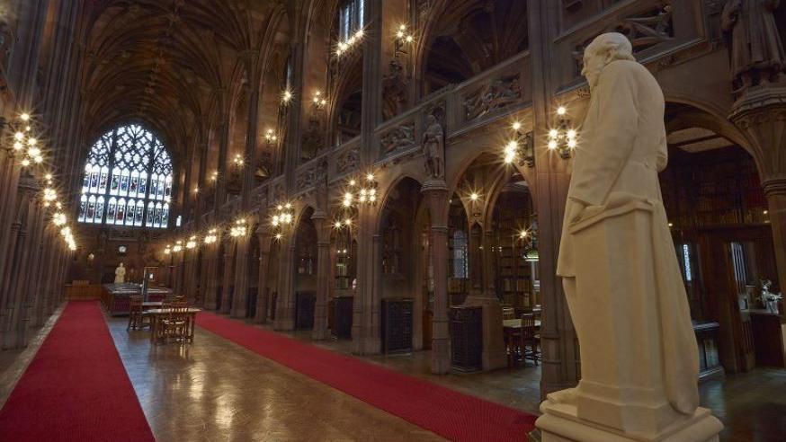 Main hall leading to a stained glass arch window below a vaulted wooden ceiling. Red carpets line either side of the hall with tables in between, while a white marble statue of John Rylands looks across it from the right. On either side of the walkway are shelves of books and an elevated walkway.