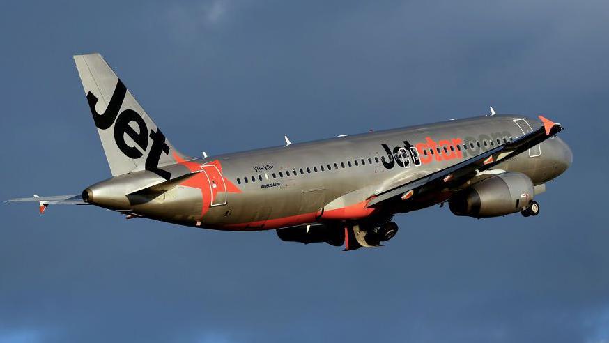 A silver plane with Jetstar's orange and black livery flies against a dark cloudy sky.