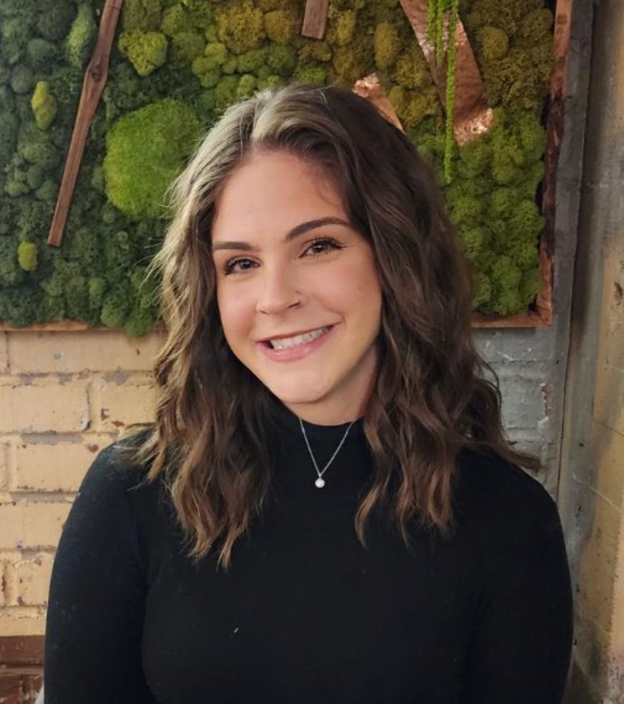 Olivia, a 33-year-old woman, wearing a black jumper. She is smiling at the camera and is sitting in front of a white brick wall with some foliage decoration