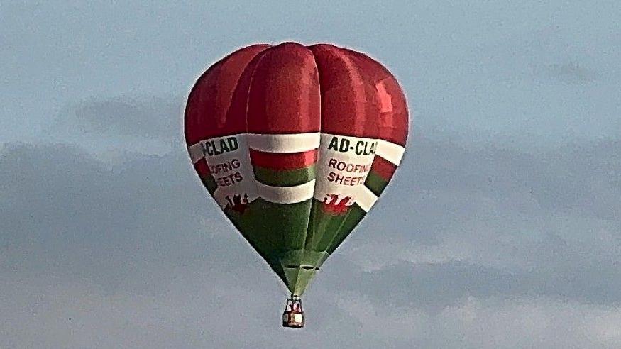 A hot air balloon in flight. It is red, white and green, with two red Welsh dragon symbols on two of the balloon's panels. The words AD-Clad Roofing Sheets are also displayed twice.