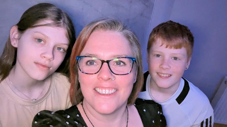 Shows a girl on the left, a woman in the middle wearing glasses with a ring through her left nostril, and a boy with red hair on the right