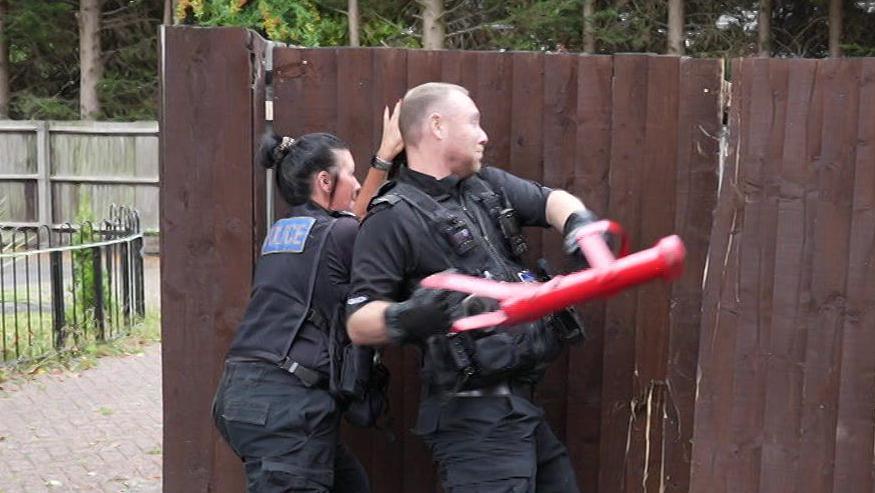 Two officers use a red "enforcer" to open a garden gate