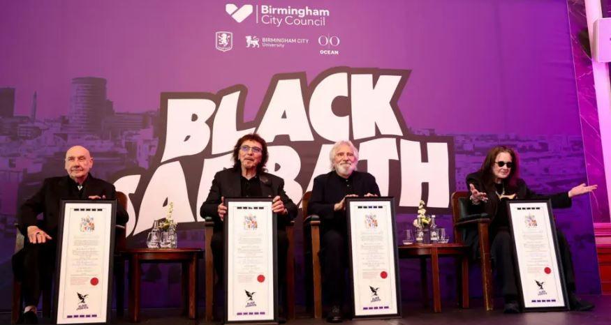 From left to right, Bill Ward, Tony Iommi, Geezer Butler and Ozzy Osbourne receive their scrolls. There is a council branded Black Sabbath board behind them in purple. They are sat in front of it on chairs. All are wearing black. Ozzy has his arms outstretched.