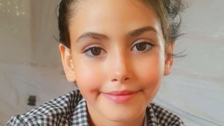 A young girl with brown eyes and dark hair, wearing a checked shirt, smiles at the camera