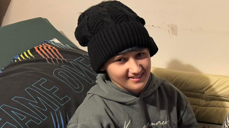 Archie sits on his bed, holding toys and other presents. He wears a black beanie hat and a grey hoodie.