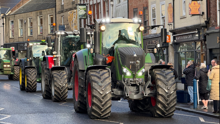 A convoy of tractors is passing through Morpeth town centre. You can see about six tractors and members of the public are watching.