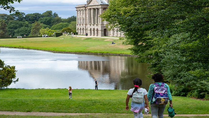 Two children with back packs walking down a hill towards a lake with two other children playing nearing the water at Lyme House in Stockport. A number of trees are in the distance.  