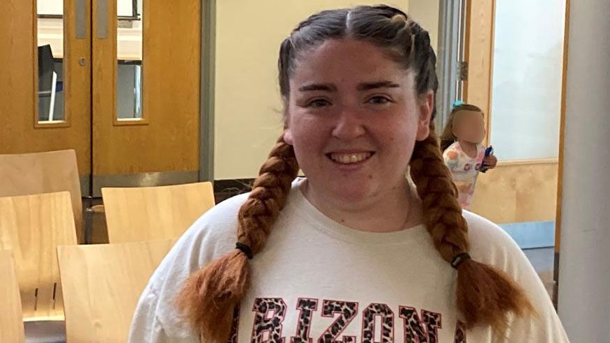 Aimee is standing in a vaccination clinic's waiting room. She has long brown hair in plaited bunches and is wearing a white jumper as she smiles for the camera.