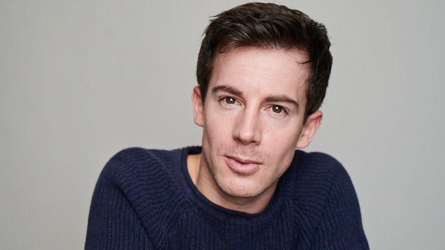Philip Labey smiling at the camera. He is wearing a navy blue knitted jumper and has brown eyes and brown short hair. The background is plain and grey.