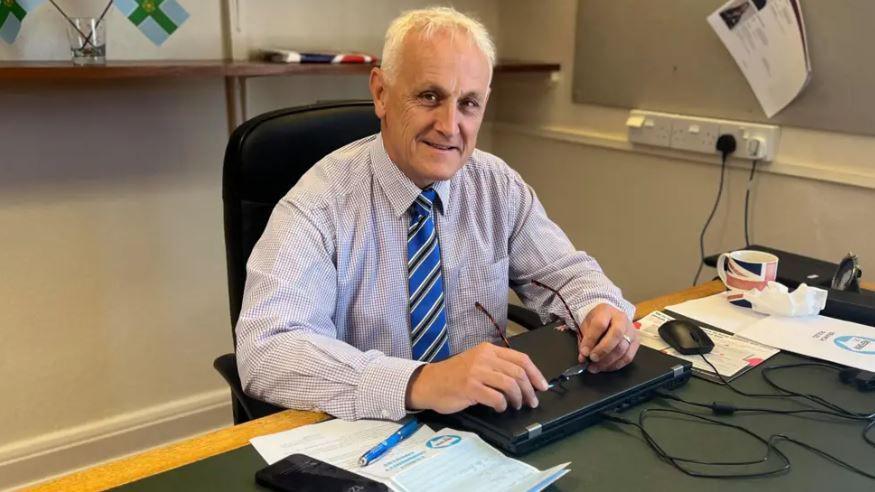 Alan Graves at a desk holding a pair glasses. He has papers, a mug and a laptop at his desk